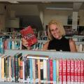 Sueli segura livro entre estantes cheias de livros. ela sorri. #paratodosverem