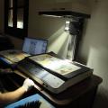 HOMEM ESTÁ SENTADO ELE USA LUVAS E MANUSEIA UM SCANNER SOBRE O QUAL HÁ UMA FOLHA DE JORNAL. #paratodosverem