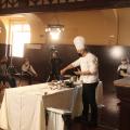 #pracegover Com uniforme de chef de cozinha, Júnior Monteiro maneja talheres sobre mesa à frente de câmeras 