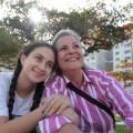 giulia entrelaça o braço direito da avó. Elas estão lado a lado, sentadas no jardim da praia. #paratodosverem. 