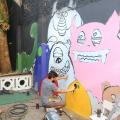 Artista, agachado, realiza pintura de monstrinho em parede de área interna da escola. #pracegover