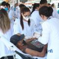 mulheres usando avental branco fazem curativo no braço de homem deitado em maca. #paratodosverem