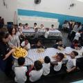 Alunos estão no refeitório tomando lanche. Todos estão sentados em torno de mesas. #Pracegover