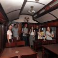 #pracegover Dentro de vagão de trem, três homens e três mulheres observam decoração e mesas de madeira do início do século 20
