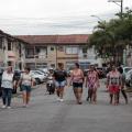 mulheres caminham por rua de uma vila. Nas laterais, os carros estão estacionados a 90 graus. #paratodosverem 