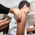 Mãos femininas aplicam injeção em braço de adolescente. O adolescente segura na mão de alguém. #Paratodosverem