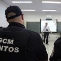dois guardas municipais com coletes, sentados, de costas para a foto. Ao fundo, aparecendo entre o meio dos ombros deles dois, há uma mulher falando à frente de um telão. #paratodosverm