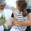 enfermeira aplica vacina no braço de criança no colo de responsável #paratodosverem