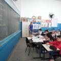 Sala de aula com professora ao fundo. A lousa está ao lado esquerdo da imagem. Os alunos estão sentados em suas carteiras. #Pracegover