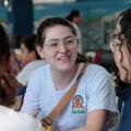 Bruna está sentada, de frente para a foto, conversando com amigas que aparecem de costas para a foto. #paratodosverem