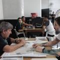 dois homens sentados frente a frente com uma mesa no meio. Um deles entrega documentos. #paratodosverem