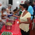 mesa com travessas fundas com comida. De um lado da mesa, pessoas servem. Do outro, são servidas. #paratodosverem