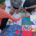 jovens estão sentados no chão, com pinceis. Eles pintam sobre tecidos. #paratodosverem