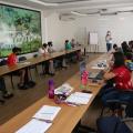 Jovens estão sentados à mesas colocadas lado a lado. Eles olham para a mulher que está ao fundo da sala, que está diante de uma projeção na parede. #paratodosverem