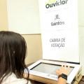 Aluna está de costas para imagem. Ela digita em uma urna eletrônica. À frente se lê em um painel: Eleição Aluno Ouvidor, Cabine de  Votação. #Paratodosverem