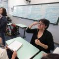 interprete de libras está na frente de aluno se comunicando. Ao fundo, a professora dá aula para a classe. #paratodosverem