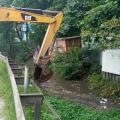 Trator com escavadeira realiza limpeza em córrego ao lado de área verde #paratodosverem