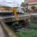 escavadeira retira mato da superfície de canal. Homem está sobre um cano dentro do canal, fazendo a retirada manual de resíduos. #paratodosverem