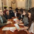 Autores, sentados, conversam com estudantes, em pé, que observam o livro. #paratodosverem