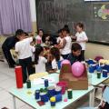Onze crianças se reúnem em mesa com objetos reciclados. À frente, professora mostra objeto a aluna, junto a mesa com materiais coloridos