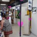 Corredor da escola está decorado com exposição de fotos e corações cor de rosa. À esquerda da imagem há uma mulher e uma criança olhando fotos que estão sobre uma mesa. #Pracegover