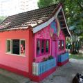 Casa de boneca com fachada frontal e lateral à vista. Lateral tem duas janelas abertas. Na frente, há duas janelas, porta e dois cercadinhos para plantas. Na frente há decoração com motivos infantis.  #Pracegover