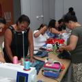 mulheres estão trabalhando em costura em torno de uma mesa. Em primeiro plano está uma máquina de costura. #paratodosverem