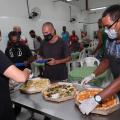 Uma pessoa atrás de uma mesa , onde estão três pizzas , algumas pessoas na fila para comer #paratodosverem