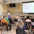 salão com pessoas sentadas assistindo uma palestra. Ao fundo uma mulher em pé fala ao grupo. #paratodosverem