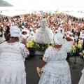 mulheres vestidas de branco estão em palco de costas para a foto. Imagem de iemanjá está à frente com o público ao fundo. #paratodosverem