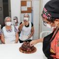professora decora bolo com brigadeiros. Ao fundo, mulheres assistem à aula. Uma delas fotografa com o celular. #paratodosverem