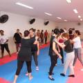 mulheres estão em pé sobre tatame. Algumas estão em dupla fazendo movimentos de defesa. #paratodosverem