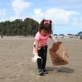 criança usa luva para pegar um microlixo na faixa de areia. Ela segura um saco de papel para depositar o resíduo.#paratodosverem