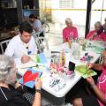 idosos estão sentados em torno de uma mesa pintando telas. A mesa está cheia de apetrechos como tintas e pinceis. #paratodosverem 