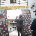 homem em primeiro plano, de costas, olhando para dentro de uma sala em cuja entrada estão afixadas inúmeras fotos. Outro homem fotografa a sala com celular. #paratodosverem