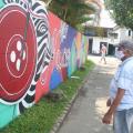 homem em pé na praça usando máscara olha para pintura em muro. . Trecho da calçadaé ajardinado. #paratodosverem