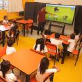 professor está diante de tela apontando para figura e crianças estão sentadas prestando atenção. #paratodosverem