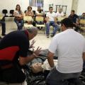 dois homens ajoelhados no chão simulam atendimento a  pessoa ferida. Trata-se de um boneco deitado também no chão. Ao fundo, os alunos atentos. #paratodosverem