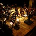 Músicos da orquestra tocam em palco com maestro à frente. #paratodosverem
