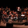 maestro está à frente da orquestra. Ele olha para a frente. Músicos estão atrás. #paratodosverem