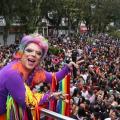Drag Queen está no alto de um carro alegórico saudando uma multidão em praça. #Pracegover