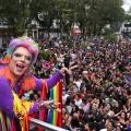 Drag com roupa colorida no alto de carro de som mostra a praça lotada ao fundo. #paratodosverem