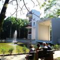 área verdade, com árvore e primeiro plano, banco com quatro pessoas ao lado. Elas estão olhando para um lago com um jato d'água ao centro, que tem, à direita, um mirante de três pavimentos. #paratodosverem