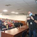palestrante fala no microfone com público assistindo ao fundo #paratodosverem