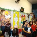 Professora, pais e alunos reunidos em uma sala de educação infantil. A educadora está fazendo uma apresentação aos pais. A parede da sala tem várias colagens de  motivos infantis. #Pracegover