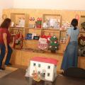 duas mulheres mexem em objetos artesanais expostos em prateleiras. Elas estão de costas para a foto. Em primeiro plano, uma miniatura da casa do trêm bélico sobre uma mesa. #paratodosverem 