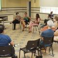 várias pessoas sentadas em roda em uma sala. Uma delas, ao fundo, está falando. #paratodosverem