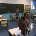 sala de aula com crianças sentadas, todas de costas. A professora está ao fundo, à frente da lousa e falando aos alunos. #paratodosverem