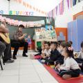 O músico está sentado tocando violão. Há uma professora ao lado dele. As crianças estão sentadas  no chão e acompanham. #Pracegover