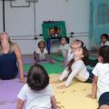 Crianças e professora fazem movimento no solo sobre tapetes de EVA. Elas estão com o corpo no solo e têm os dois braços esticados e paralelos. Todos flexionam a cabeça e olham para o alto. #Pracegover